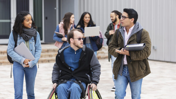 A multiracial group of young adults, university students, hanging out outside a campus building, conversing. The friends include a man with cerebral palsy in a wheelchair. Three of the students are walking and conversing in the foreground while three others are out of focus in the background.