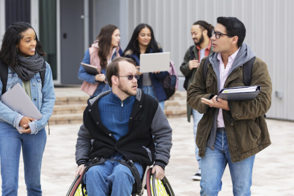 A multiracial group of young adults, university students, hanging out outside a campus building, conversing. The friends include a man with cerebral palsy in a wheelchair. Three of the students are walking and conversing in the foreground while three others are out of focus in the background.