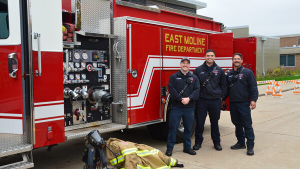 Three fire fighters stand in front of a red fire truck outside near a pile of protective gear.