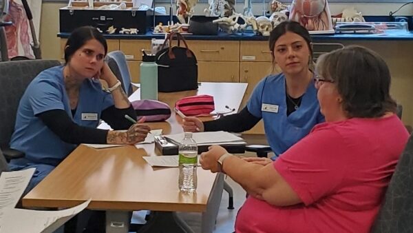 2 students in scrubs sitting at table interviewing mock patient