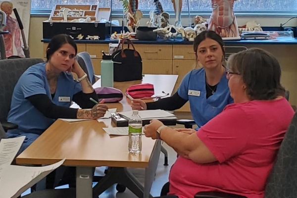 2 students in scrubs sitting at table interviewing mock patient