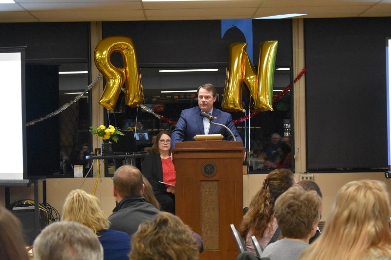 man at podium with R and N balloons behind him and an audience in front of him