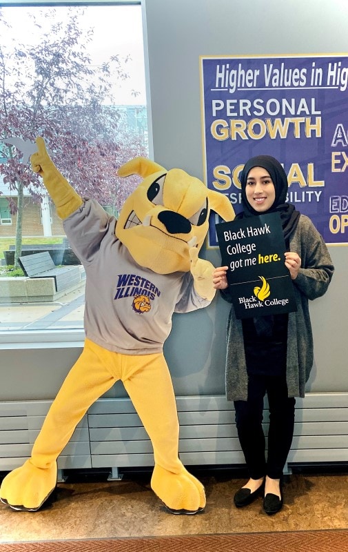 student standing next to a bulldog mascot while holding a sign that says Black Hawk College got me here