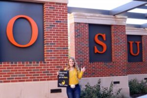 Grayson Cottrell standing in front of Oklahoma State University building, holding sign that says "BHC got me here"