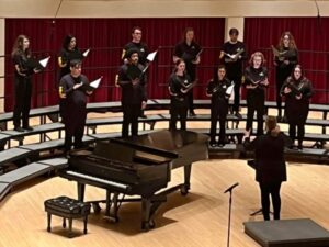 Choir director on stage with 12 choir members on risers singing