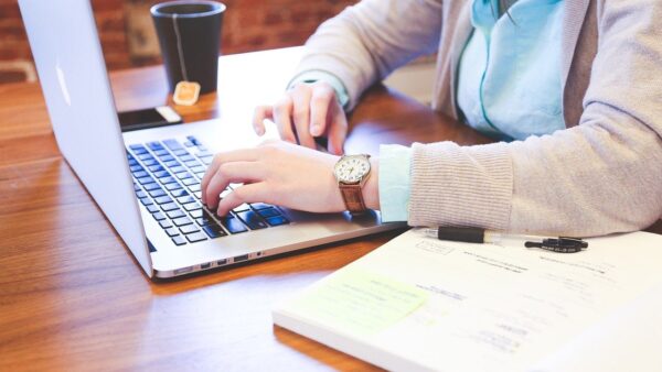 hands typing on computer next to book on table