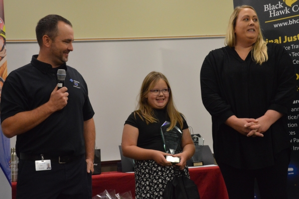 adult holding microphone standing next to girl who is holding award and her mother