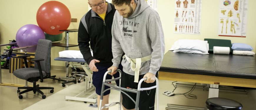 A student stands beside a man using a walker in a physical therapy setting