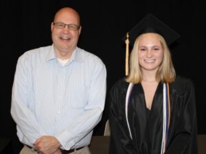 2 people standing in front of black backdrop with one wearing graduation cap & gown