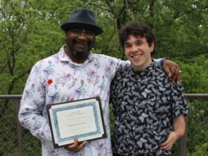 2 people standing outside with trees in background and one person holding a certificate