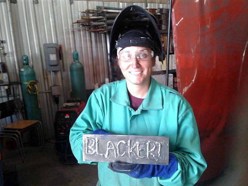 smiling woman in welding gear holding a welded plaque that says her last name, BLACKERT