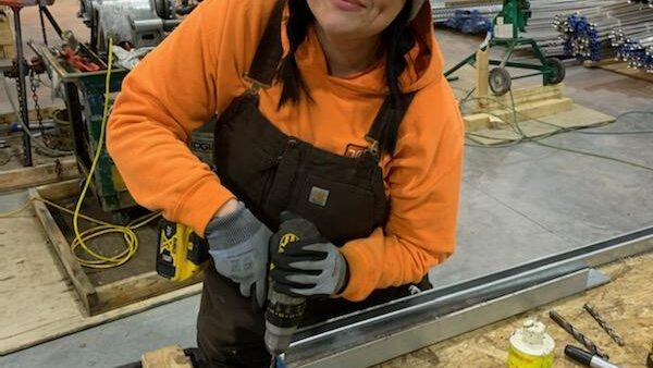 woman wearing safety glasses and a hard hat smiles while holding a drill at a work station