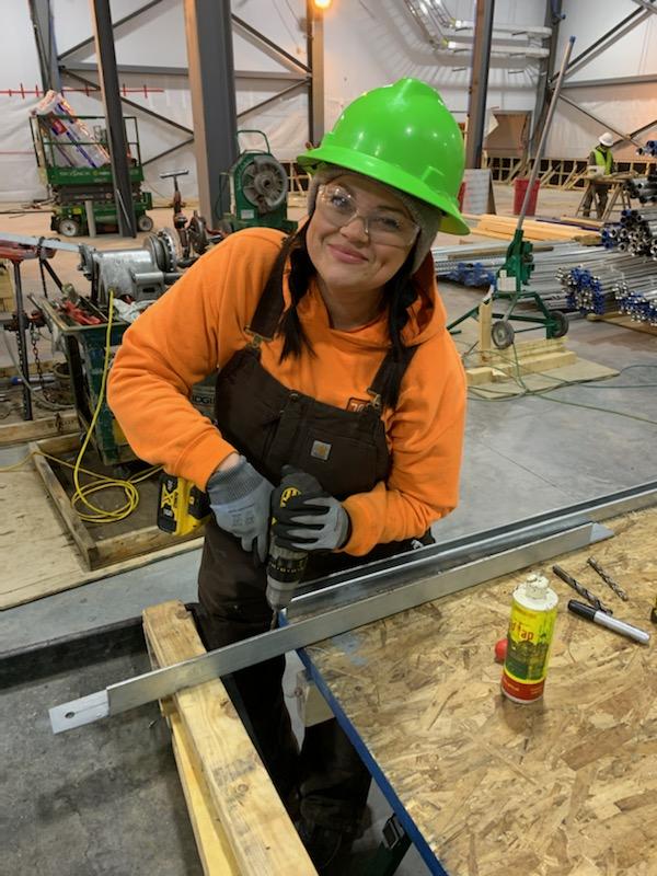 woman wearing safety glasses and a hard hat smiles while holding a drill at a work station