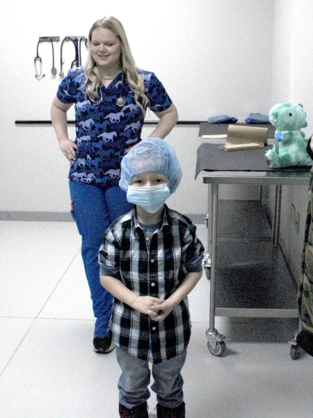 child wearing surgical mask and cap with stuffed animal and college student wearing scrubs in background