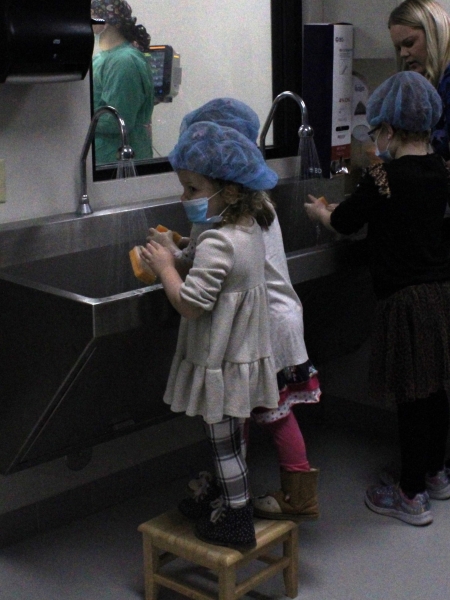 3 children wearing surgical masks and caps scrubbing hands at sink