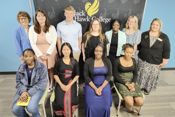 7 people standing and 4 people sitting in front of Black Hawk College logo backdrop