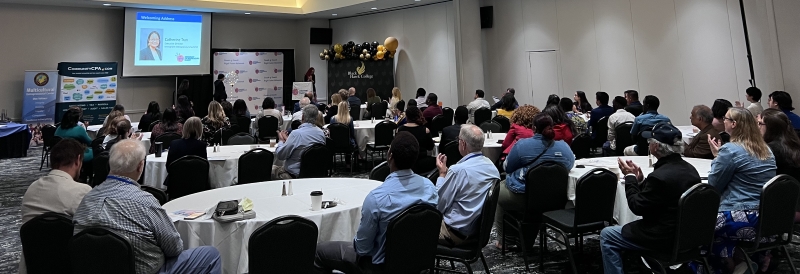 large room full of people at tables and speaker at podium in front of screen