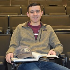 student sitting in theater room with book open on lap and hat resting on book