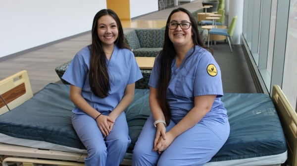 2 student nurses wearing scrubs sitting on hospital bed