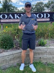 Luke Lofgren standing in front of Oakwood Country Club sign with 2 awards