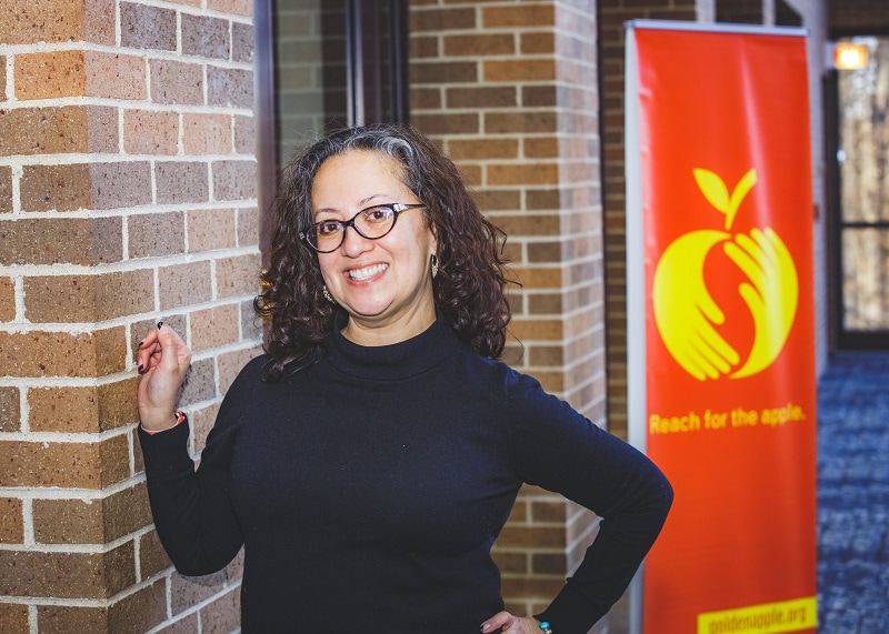 teacher standing next to brick wall with a red banner in the background featuring a logo of hands creating an apple