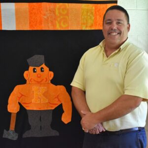 Kewanee High School teacher standing in front of a quilt featuring the school's mascot, a boilermaker