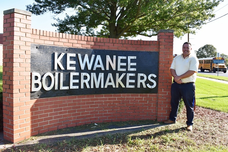 teacher standing next to a brick Kewanee Boilermakers sign with a school bus in the background