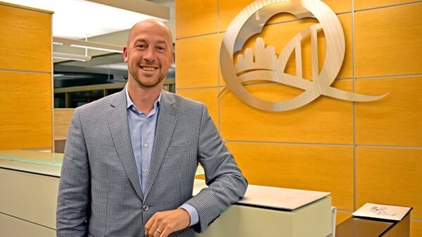 man in suit in front of a desk with a Q-shaped logo on the wall featuring a cityscape