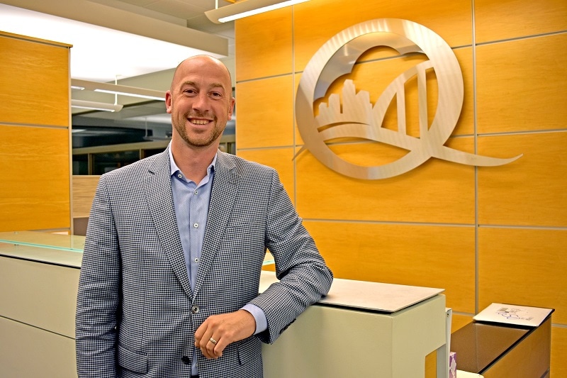 man in suit in front of a desk with a Q-shaped logo on the wall featuring a cityscape