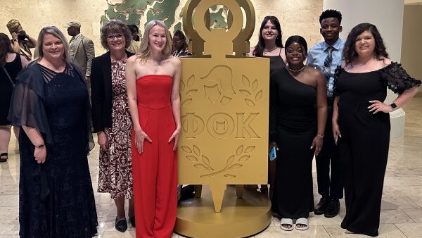 7 people dressed up & standing by large Phi Theta Kappa gold key emblem