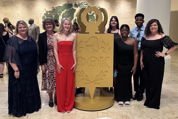 7 people dressed up & standing by large Phi Theta Kappa gold key emblem