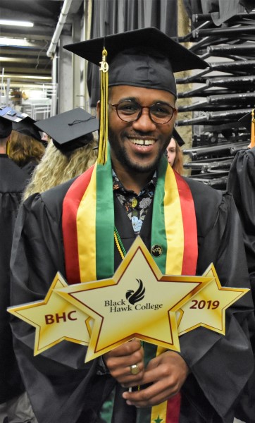 Amadou Ndiaye wearing graduation gown. Holding 3 signs shaped like gold stars saying "BHC", "Black Hawk College", and "2019"