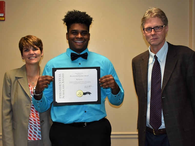 male college student holding up a certificate standing with two college officials