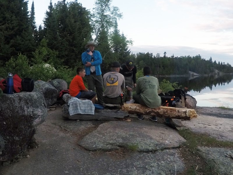 a group of scouts around a campfire on the shore of a lake