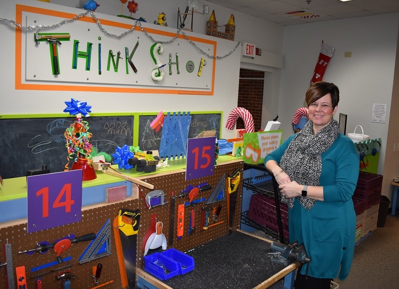 Woman leans against safety goggles rack next to work stations. ThinkShop is spelled out with tools and construction materials on the wall above the work stations.