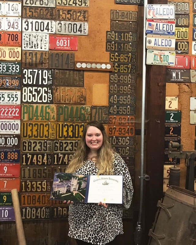 woman holding her degree in front of a wall of license plates