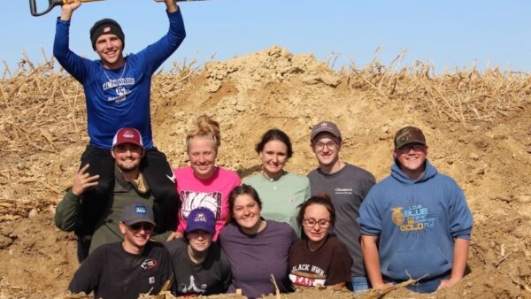 10 students in soil pit with one holding shovel above head