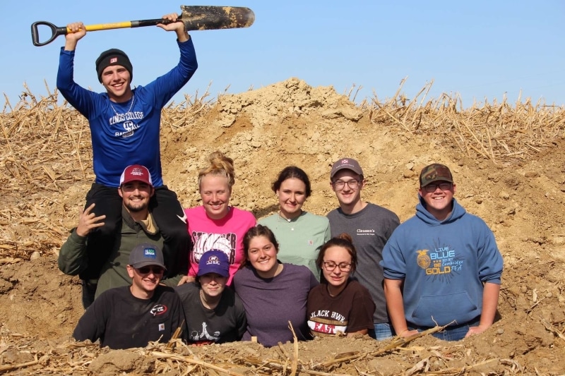 10 students in soil pit with one holding shovel above head