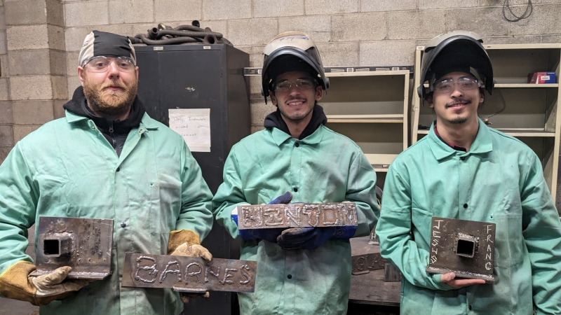 3 students in welding gear holding up nameplates they welded