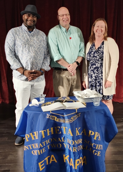 3 people standing on stage in front of small draped with Phi Theta Kappa banner