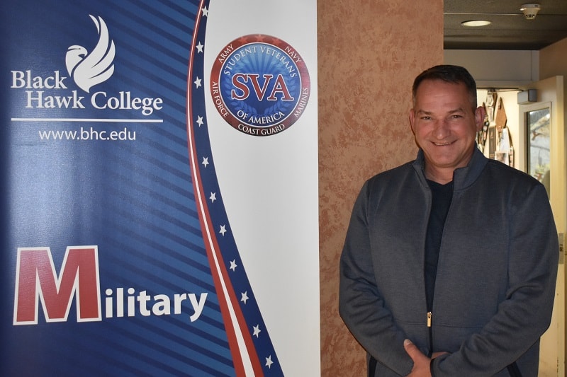 Black Hawk College Veteran's Coordinator Thomas Reagan standing next to a red, white, and blue banner for the Military Students and Veterans Club