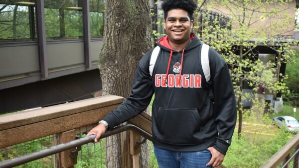 smiling male college student standing at the top of a staircase on campus with trees in the background